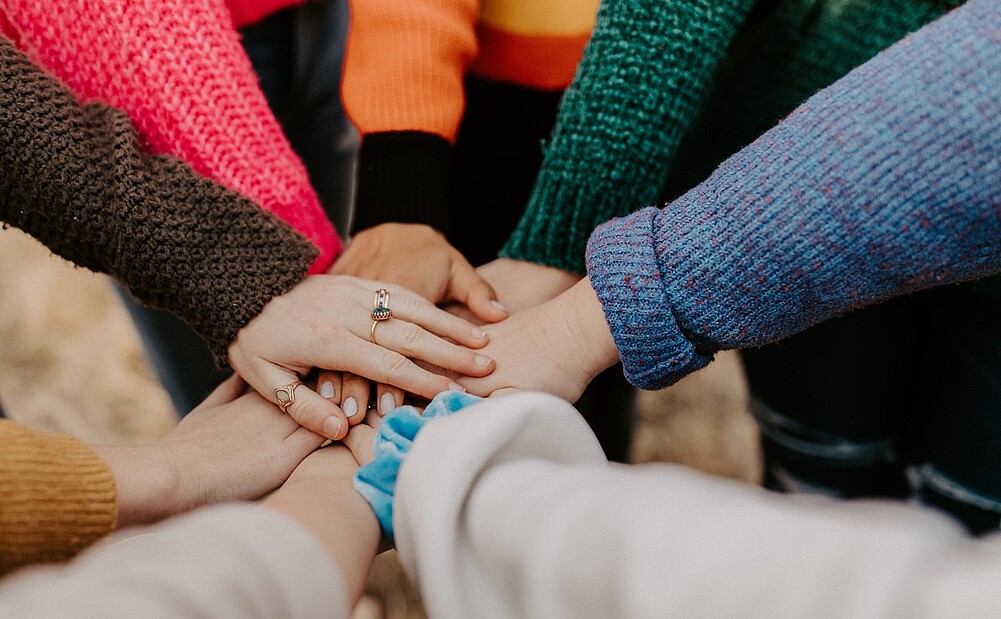 Image of a group of students standing in a circle, putting their hands together. The image represents the power of teamwork and collaboration to make positive change.