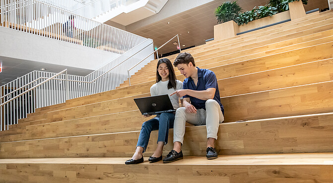 Two International Business Administration students, one holding a laptop and the other pointing towards the laptop, whilst sitting on the first floor staircase of the Polak building located on the Erasmus University campus