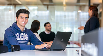 Enthusiastic Rotterdam School of Management student engaged in a workshop, wearing a cheerful smile, while diligently working on his laptop.